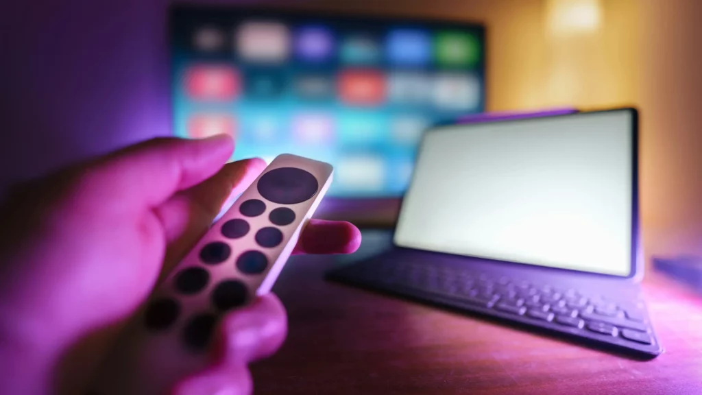 A person holding a remote control in front of a laptop. Themes of multiple mediums.