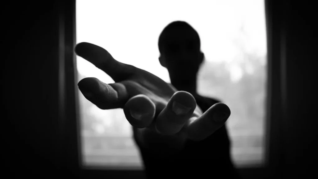 A black and white photo of a figure in shadow reaching towards the camera with an outstretched hand.