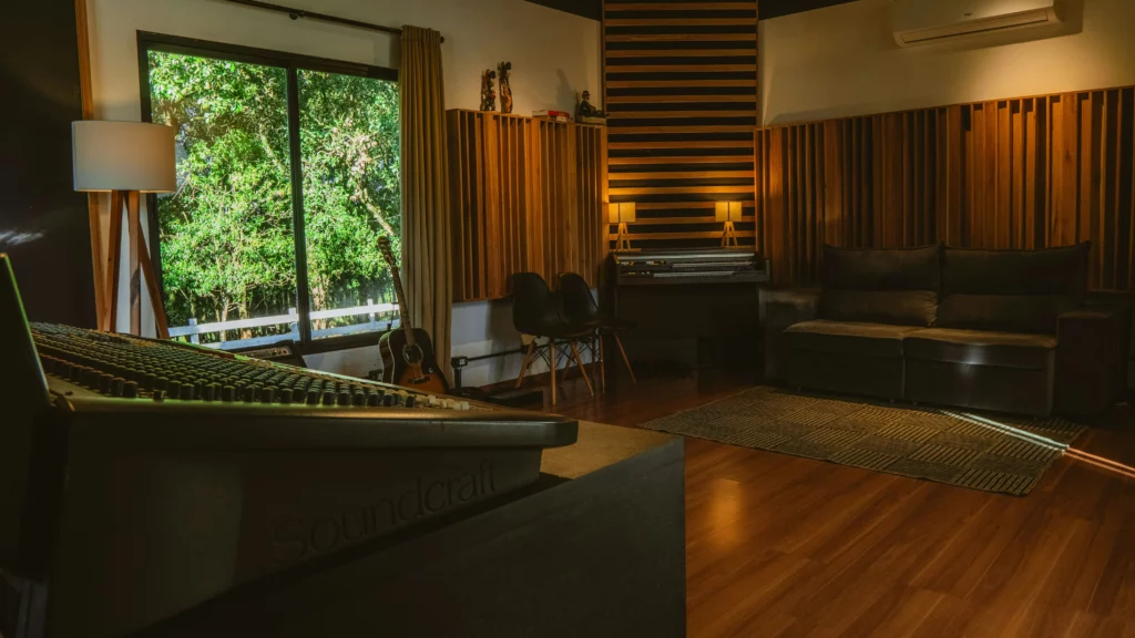 Music studio with wooden acoustic panels, mixing console, keyboard, and windows overlooking greenery.